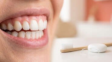 Close-up of a smiling mouth with clean, white teeth; a toothbrush and dental floss on a white surface nearby.